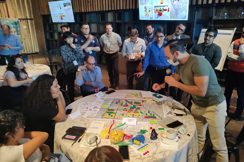 People sit and stand around a round table while a man talks into a microphone