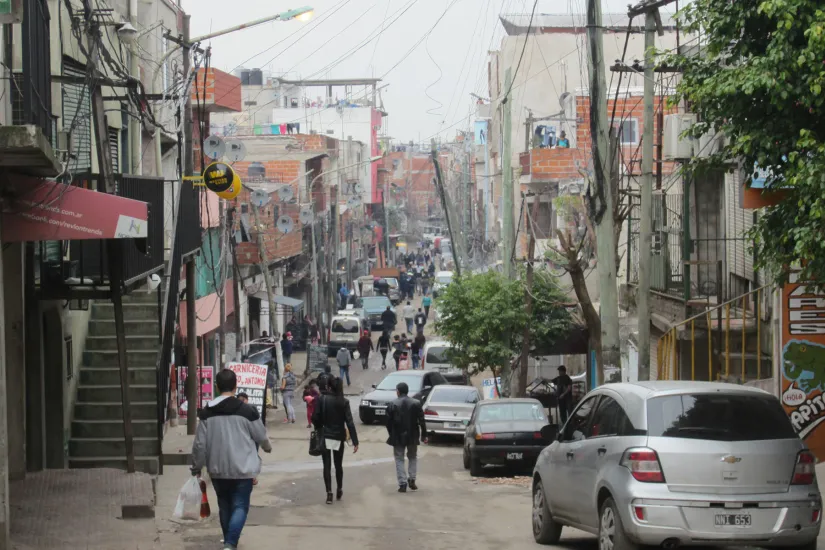 Peatones caminando por una calle con coches aparcados a los lados en Villa 20, Buenos Aires.