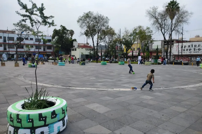 Dos niños pequeños juegan al fútbol en una plaza con arbolitos plantados en neumáticos pintados.