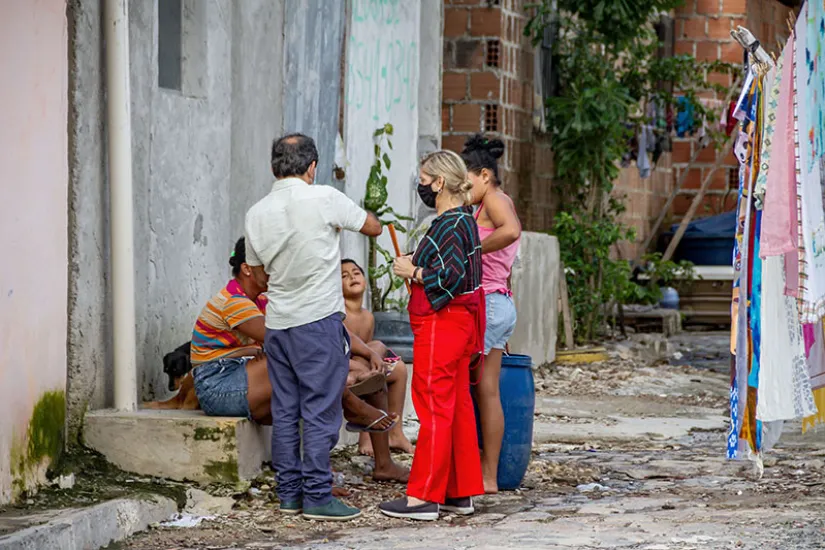 Uma mulher e algumas crianças estão sentadas na soleira de uma porta e conversam com um grupo de três pessoas.