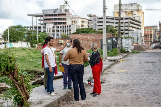 Cinco pessoas conversam em uma calçada diante de um campo com prédios brancos ao fundo.