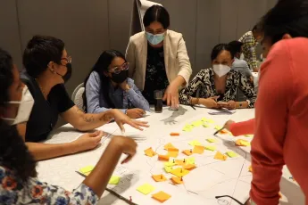 Miniatura de vídeo mostrando pessoas usando máscaras colando notas adesivas em um mapa disposto sobre uma mesa.