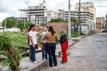 Five people talk on a sidewalk in front of a field with white buildings in the background