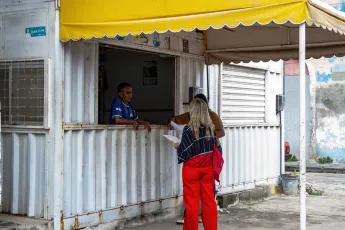 Duas pessoas estão em pé debaixo de um toldo de plástico e conversam com um homem em um container que foi transformado em moradia.
